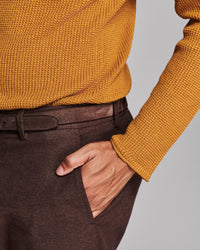 Close-up of a person wearing a mustard yellow ribbed knit sweater and dark brown trousers with a matching belt, with one hand in the trouser pocket, against a light gray background.