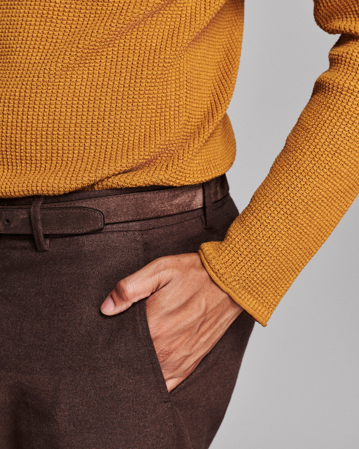 Close-up of a person wearing a mustard yellow ribbed knit sweater and dark brown trousers with a matching belt, with one hand in the trouser pocket, against a light gray background.