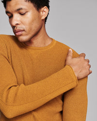 A close-up of a man wearing a textured mustard yellow knit sweater against a solid gray background.