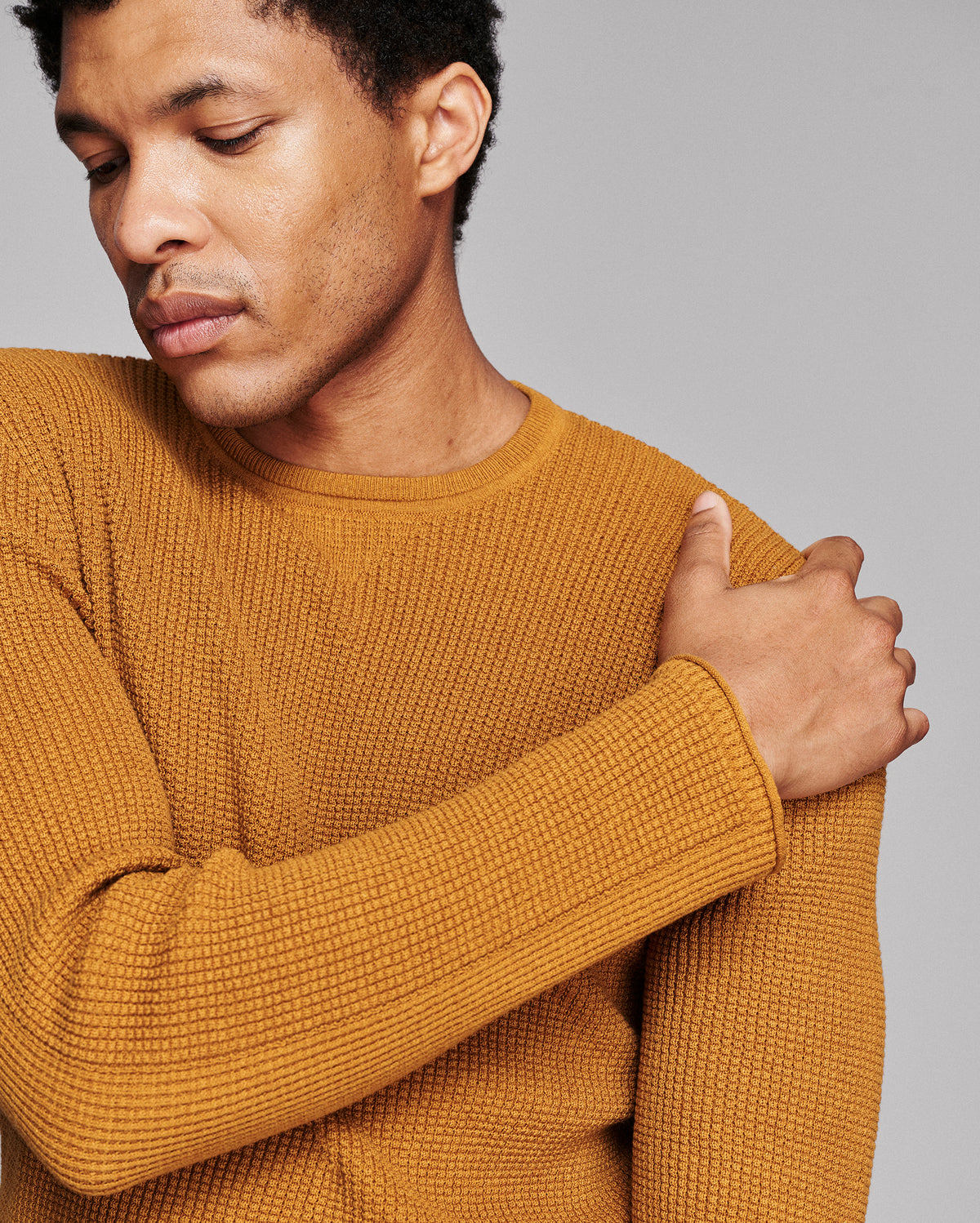 A close-up of a man wearing a textured mustard yellow knit sweater against a solid gray background.