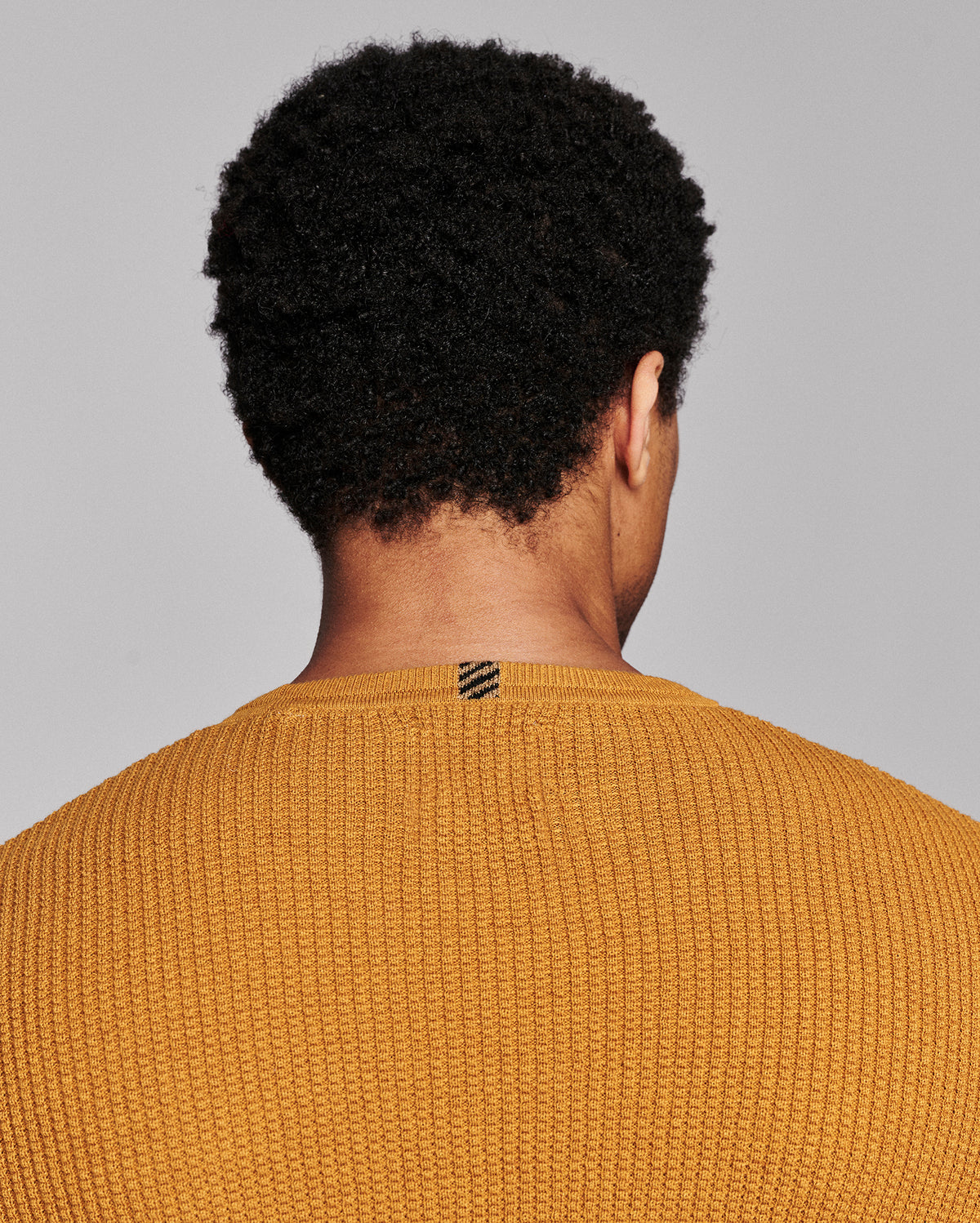 Back view of a person with curly black hair wearing a textured mustard yellow knit sweater against a solid gray background.
