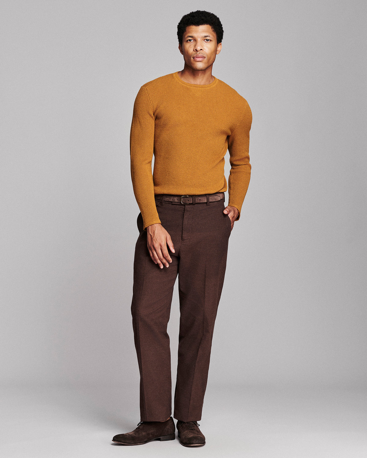 A man modeling a mustard yellow knit crewneck sweater and dark brown trousers against a light gray studio background.