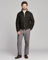 A man modeling a black zip-up jacket over a cream turtleneck sweater, paired with gray trousers and dark brown shoes, against a solid light gray studio background.