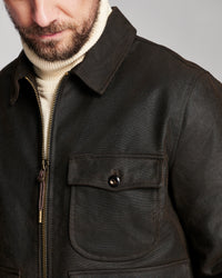 Close-up of a man wearing a dark brown waxed leather jacket over a cream turtleneck sweater, showing the collar, zipper, and chest pocket details.