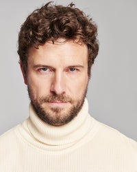 A close-up portrait of a man with curly brown hair and a beard, wearing a cream-colored ribbed turtleneck sweater against a solid light gray background.