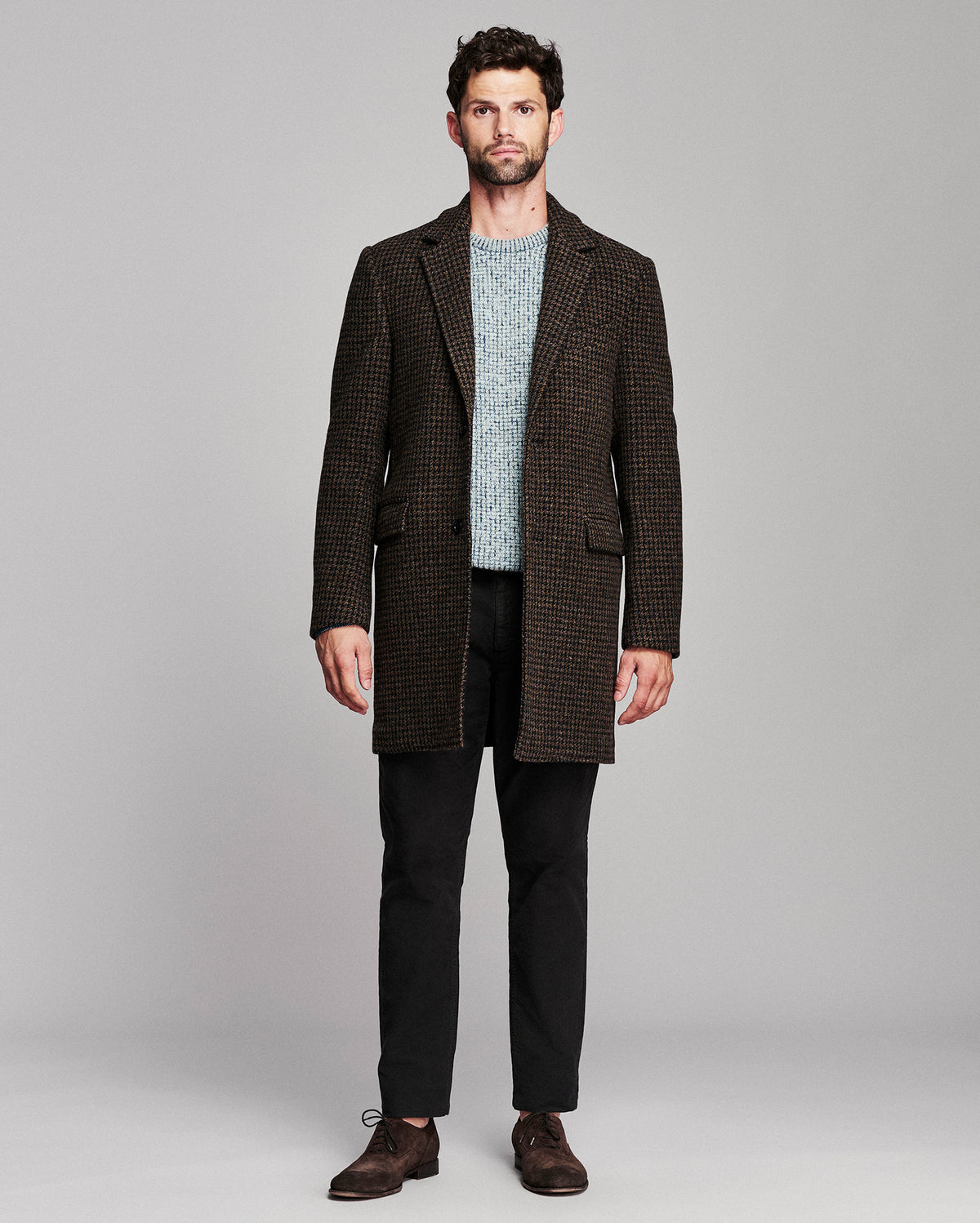 A man modeling a dark brown and black micro-check blazer over a light blue knit sweater, black trousers, and brown leather shoes, shot against a solid gray studio background.