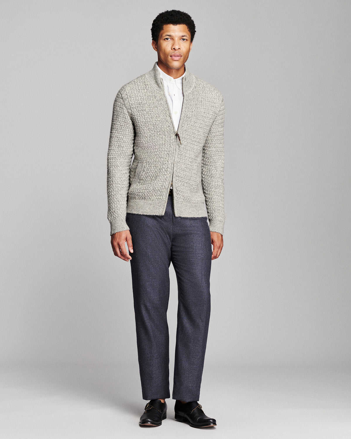A man modeling a light gray textured knit cardigan over a white collared shirt, paired with dark blue trousers and black loafers, against a solid gray studio background.