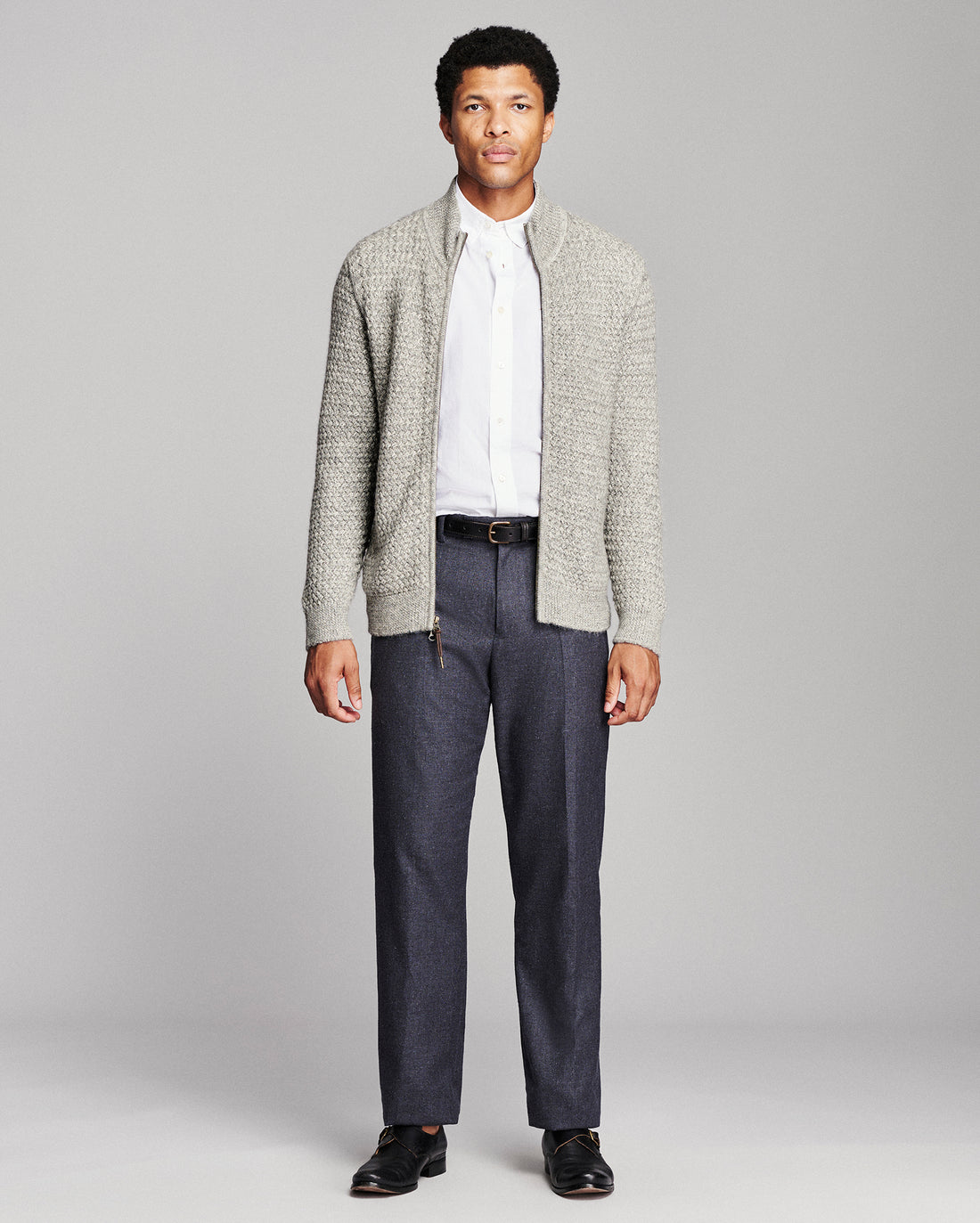 A man modeling a light gray textured knit cardigan over a white collared shirt, paired with dark gray trousers and black dress shoes, against a solid light gray studio background.