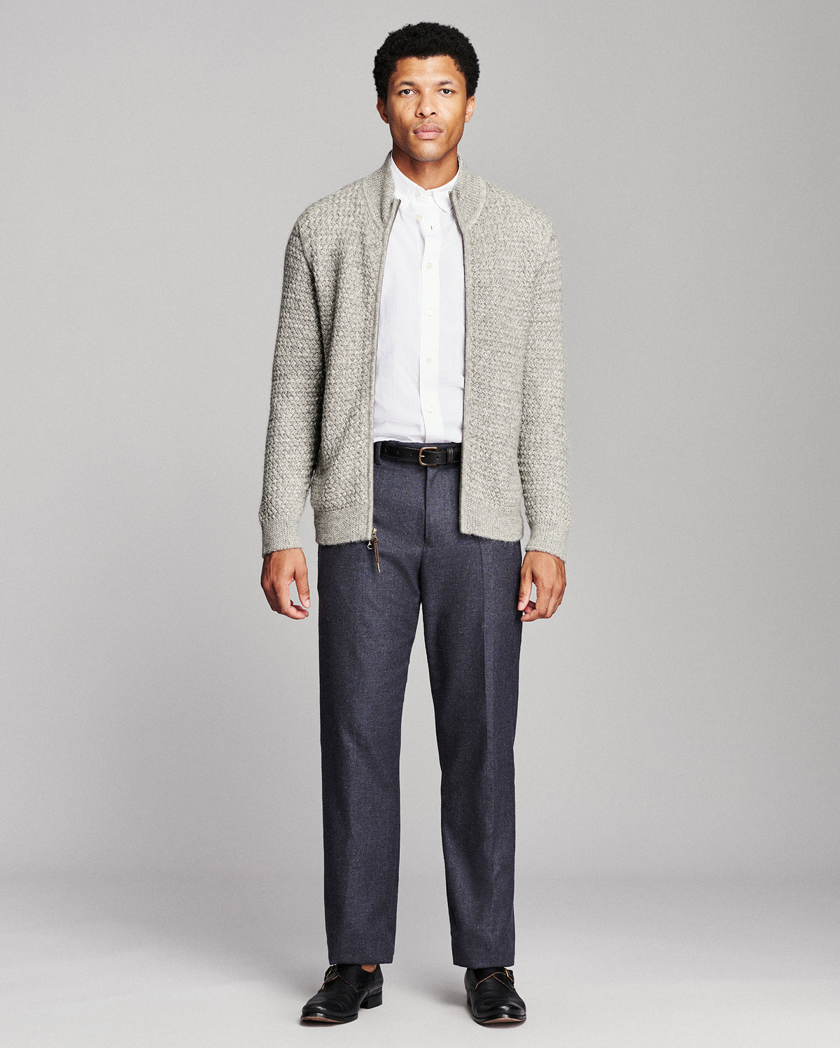 A man modeling a light gray textured knit cardigan over a white collared shirt, paired with dark gray trousers and black dress shoes, against a solid light gray studio background.