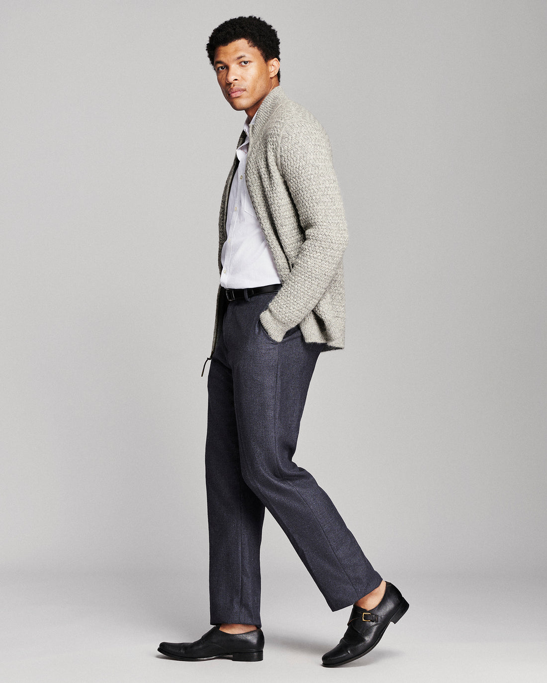 A man modeling a light gray knit cardigan over a white shirt, paired with dark gray trousers and black leather loafers, against a solid light gray studio background.