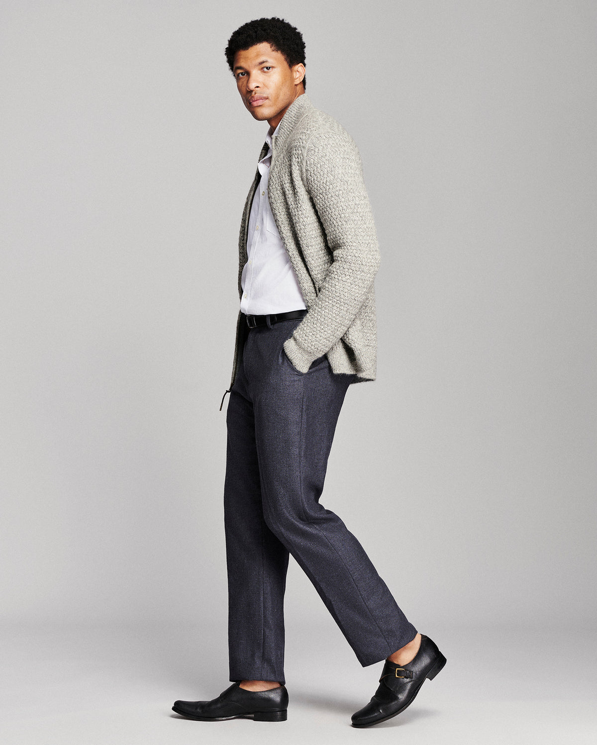 A man modeling a light gray knit cardigan over a white shirt, paired with dark gray trousers and black leather loafers, against a solid light gray studio background.