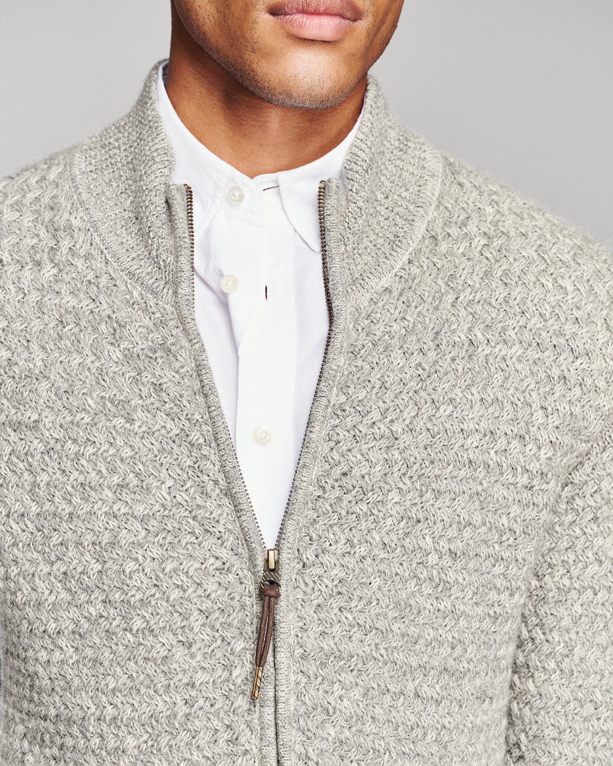 Close-up of a man wearing a light gray textured zip-up sweater over a white collared shirt, against a plain light gray background.