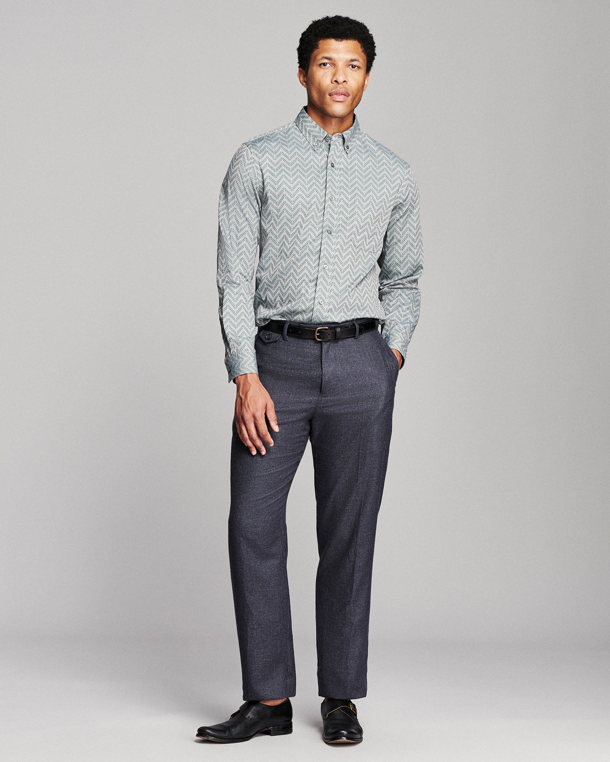 A man modeling a light gray and white chevron-patterned button-down shirt with dark gray trousers, a black belt, and black dress shoes, standing against a light gray studio background.