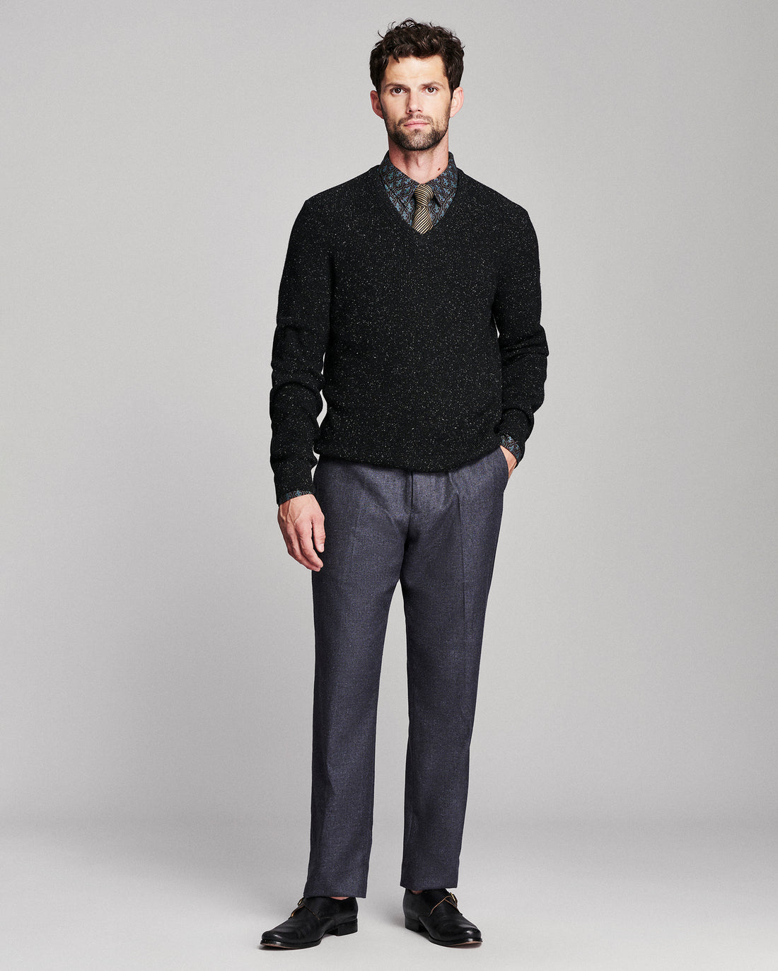 A man modeling a black speckled V-neck sweater over a patterned collared shirt, paired with charcoal gray trousers and black dress shoes, against a light gray studio background.