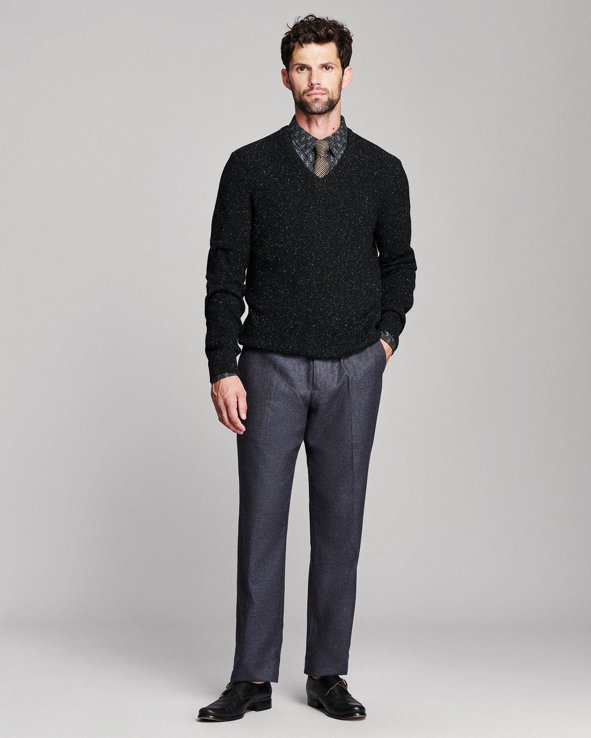 A man modeling a black speckled V-neck sweater over a patterned collared shirt, paired with charcoal gray trousers and black dress shoes, against a light gray studio background.