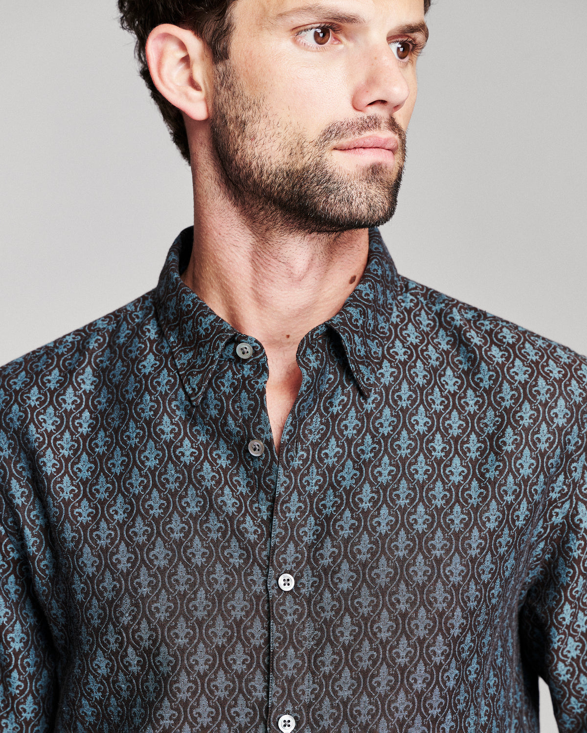 A close-up of a man wearing a dark patterned button-down shirt with a blue and brown damask-style print, photographed against a neutral gray background.