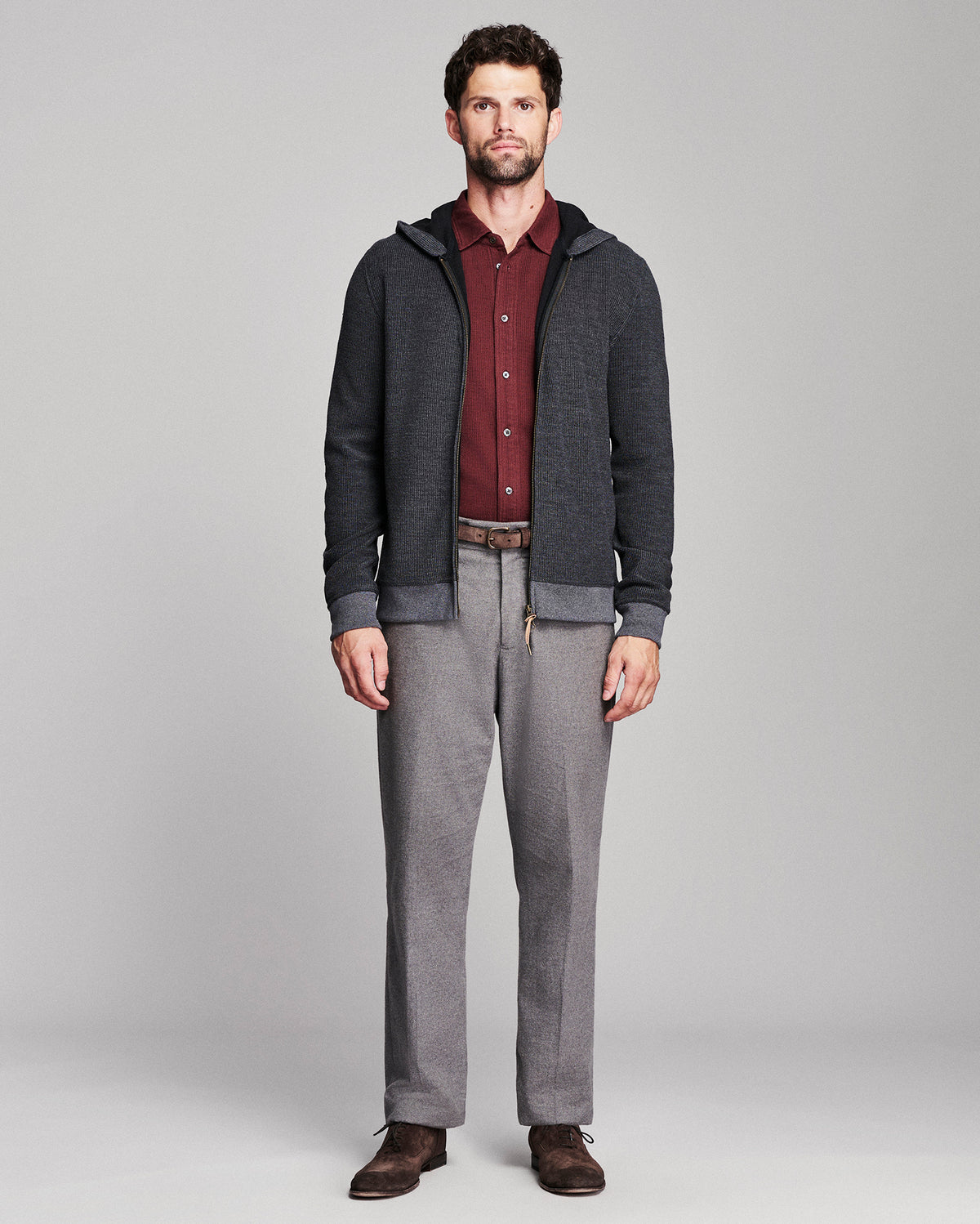 A man modeling a dark gray zip-up hoodie with a ribbed black collar, cuffs, and hem, worn over a burgundy button-down shirt and paired with gray trousers and brown shoes, against a light gray studio background.