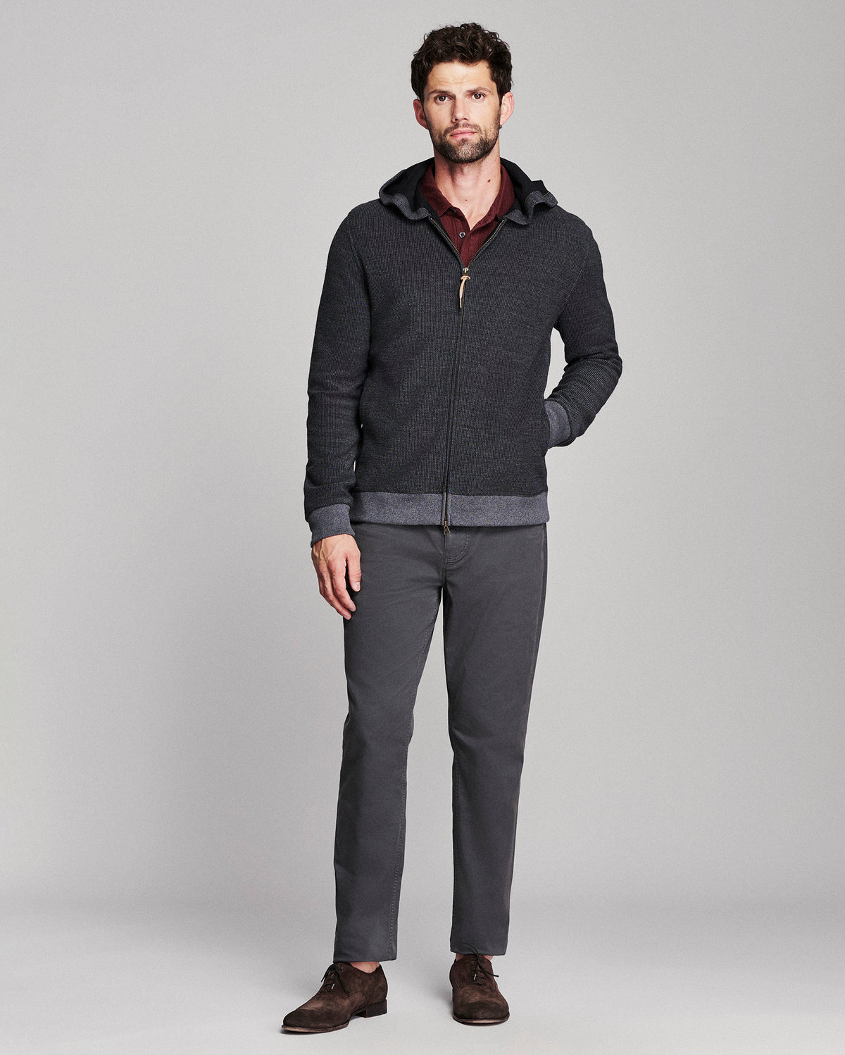 A man modeling a dark gray zip-up hoodie with a ribbed hem and cuffs, worn over a burgundy shirt and paired with dark gray trousers and brown suede shoes, against a light gray studio background.