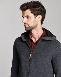 A man wearing a dark gray ribbed zip-up hoodie with a black hood and a gold zipper pull, layered over a burgundy shirt, against a solid gray background.