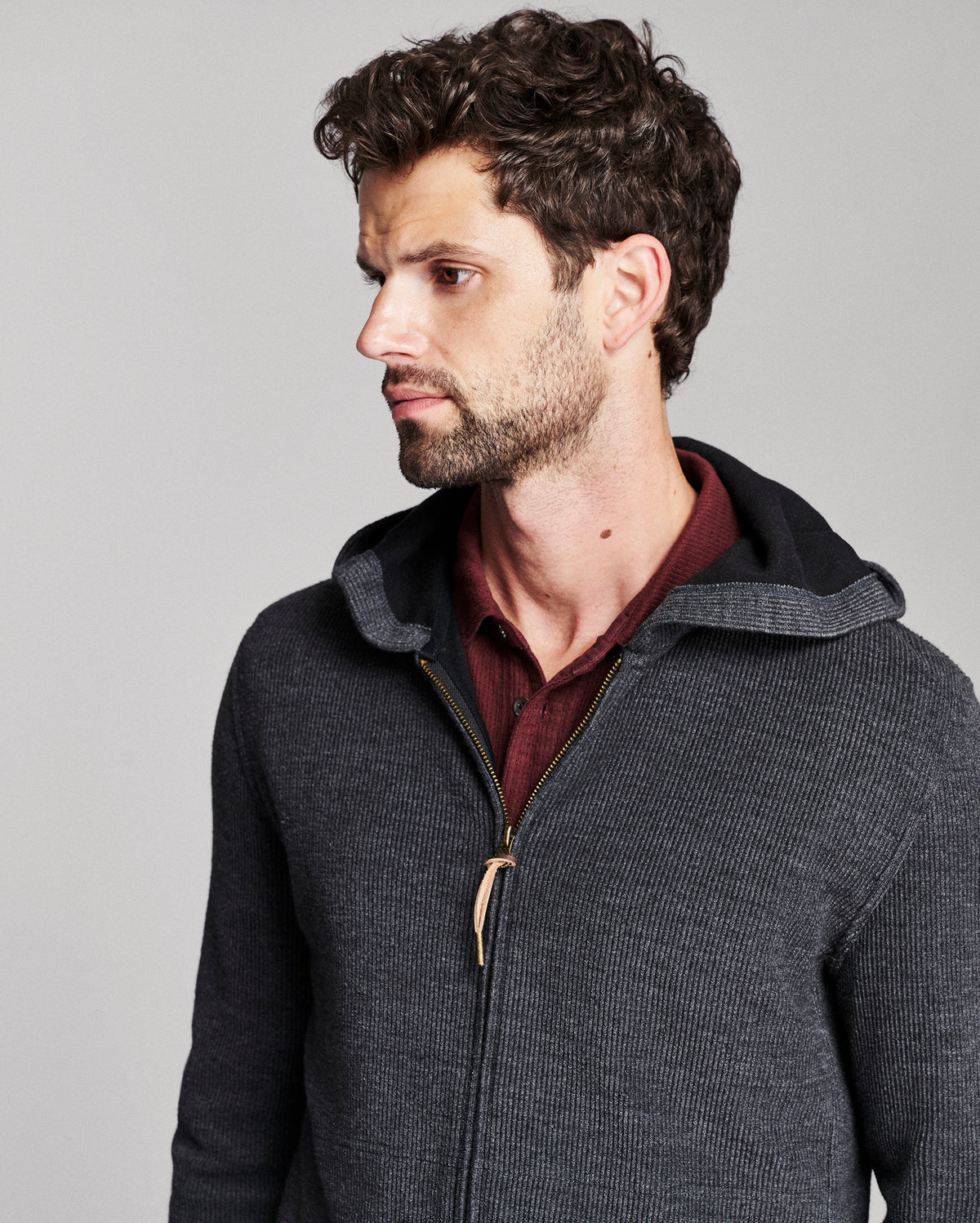 A man wearing a dark gray ribbed zip-up hoodie with a black hood and a gold zipper pull, layered over a burgundy shirt, against a solid gray background.