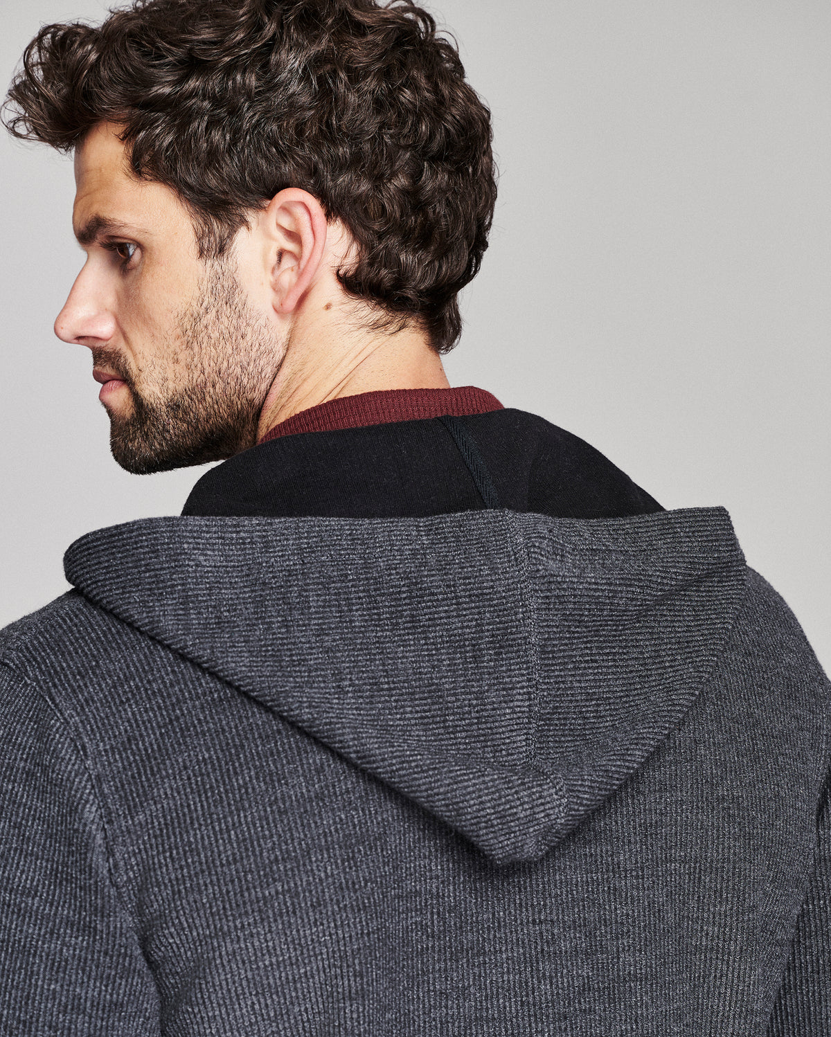 A man shown from the back wearing a dark gray textured hooded sweater over a black shirt with a burgundy collar, against a solid light gray background.