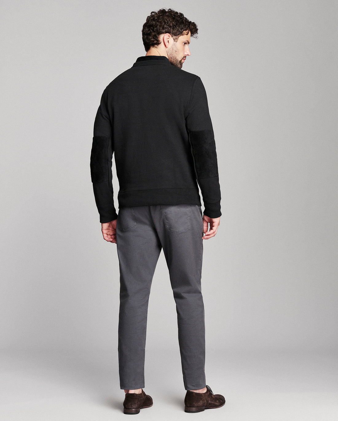 A man shown from the back wearing a black knit sweater with elbow patches, gray trousers, and dark brown shoes, photographed against a light gray studio background.