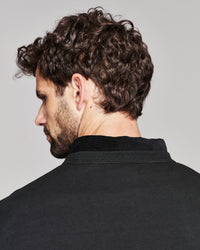 A close-up side profile of a man with curly dark brown hair and a beard, wearing a black crewneck sweater against a light gray background.