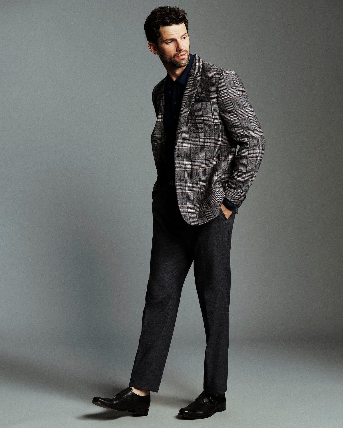 A man modeling a gray and black plaid blazer over a dark navy shirt and dark charcoal trousers, standing against a solid gray studio background.