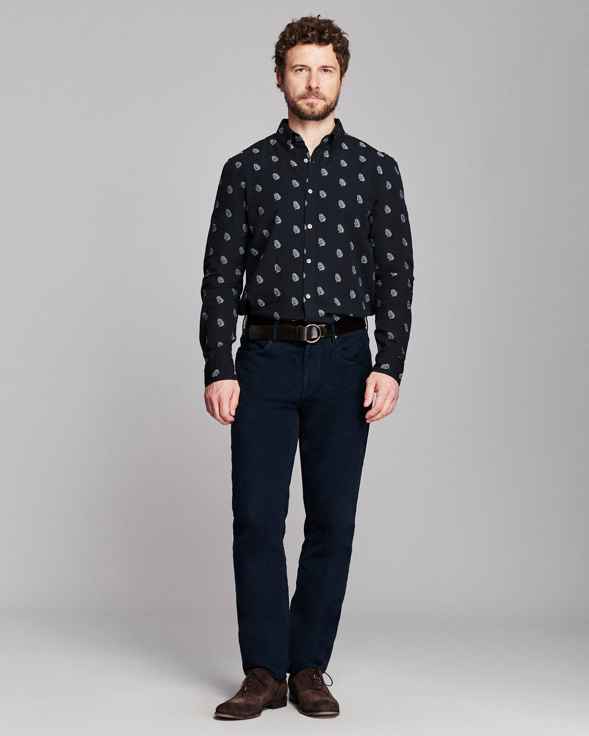 A man modeling a long-sleeve button-down shirt with a small white paisley pattern on a black background, paired with dark blue jeans, a black belt, and brown suede shoes, against a solid gray studio background.