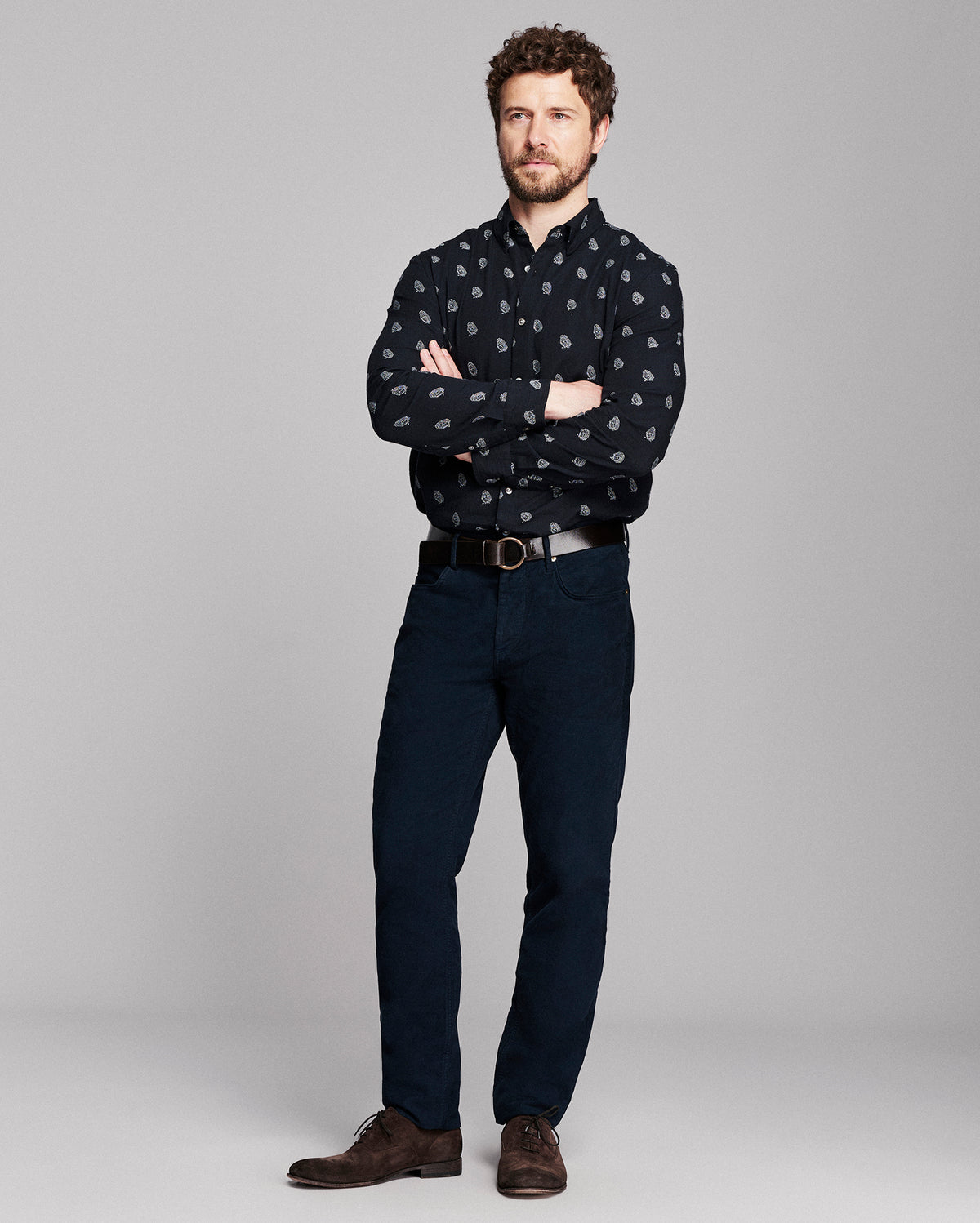 A man modeling a black long-sleeve button-down shirt with a small white paisley pattern, paired with dark blue jeans, a black belt, and brown leather shoes, against a solid gray studio background.