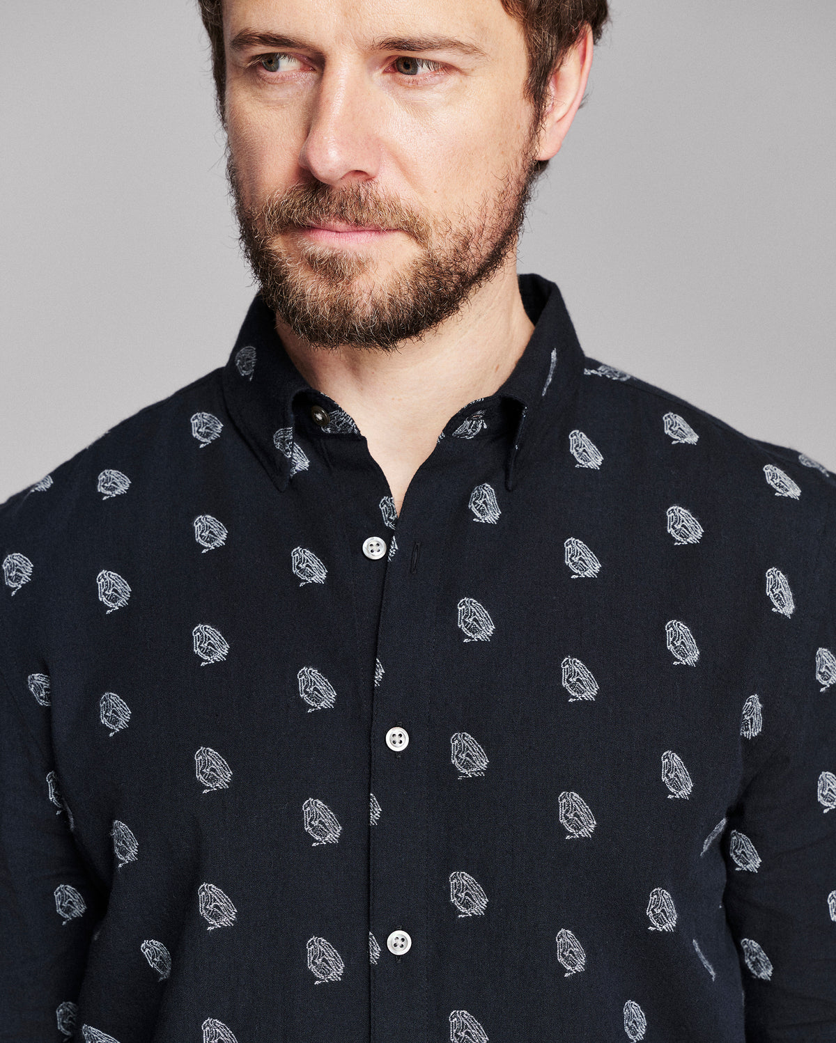A close-up of a man wearing a dark navy long-sleeve button-down shirt with a small white paisley pattern, set against a solid gray studio background.