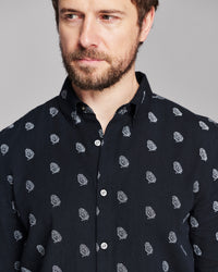 A close-up of a man wearing a dark navy long-sleeve button-down shirt with a small white paisley pattern, set against a solid gray studio background.