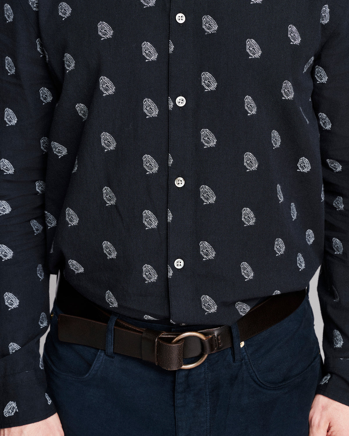 A close-up of a man wearing a dark navy long-sleeve button-up shirt with a small white paisley pattern, tucked into dark blue jeans with a brown leather belt.