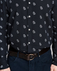 A close-up of a man wearing a dark navy long-sleeve button-up shirt with a small white paisley pattern, tucked into dark blue jeans with a brown leather belt.