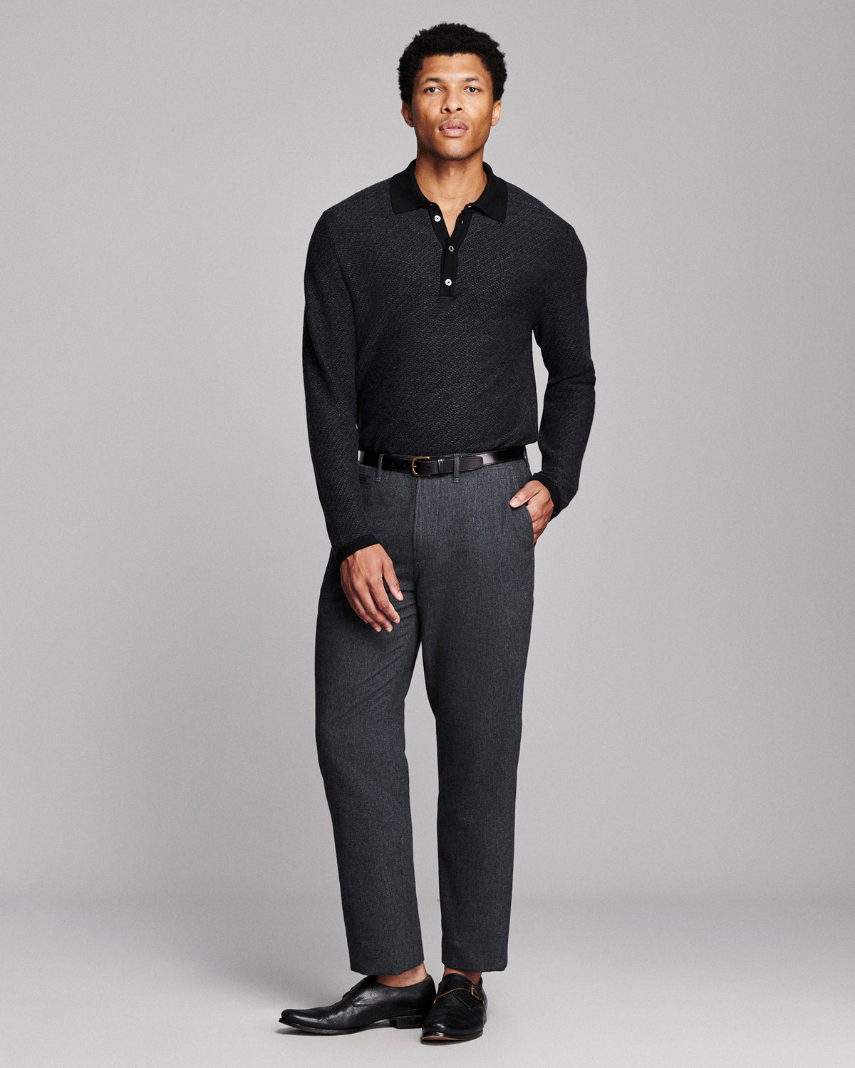 A man modeling a dark charcoal knit polo shirt and dark gray textured trousers with a black belt and black leather dress shoes, shot against a solid light gray studio background.