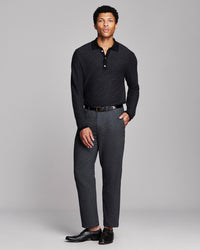 A man modeling a dark charcoal knit polo shirt and dark gray textured trousers with a black belt and black leather dress shoes, shot against a solid light gray studio background.