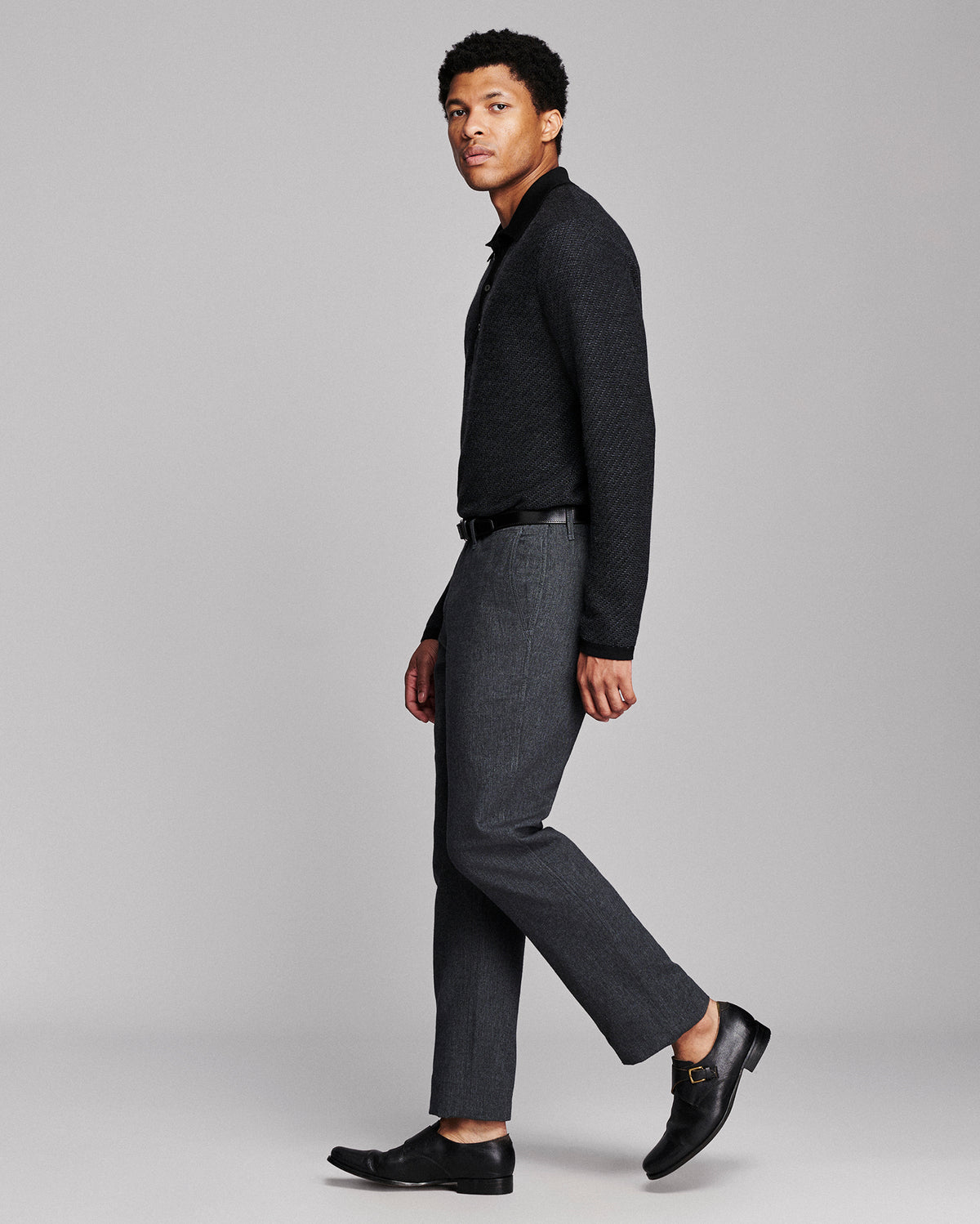 A man modeling a dark charcoal knit sweater, dark gray textured trousers, a black belt, and black leather loafers against a solid light gray studio background.