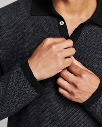 Close-up of a person wearing a dark charcoal and black textured knit polo sweater, with both hands adjusting the collar and button.