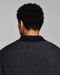 Back view of a person wearing a dark charcoal gray textured knit polo shirt with a black collar, set against a solid light gray background.
