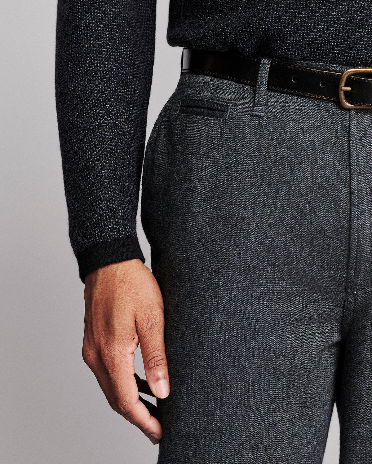 Close-up of a person wearing dark gray textured trousers, a black belt, and a dark charcoal herringbone sweater against a light gray studio background.