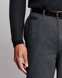 Close-up of a person wearing dark gray textured trousers, a black belt, and a dark charcoal herringbone sweater against a light gray studio background.