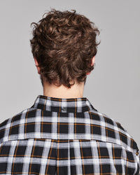 Back view of a person with curly brown hair wearing a black, white, and gray plaid button-down shirt with thin orange stripes, against a solid gray background.
