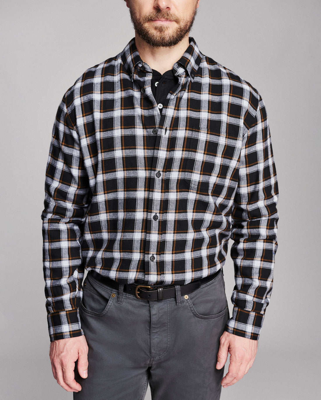 A man wearing a black, white, and thin gold plaid button-down shirt, paired with dark gray trousers and a black belt, against a solid gray studio background.