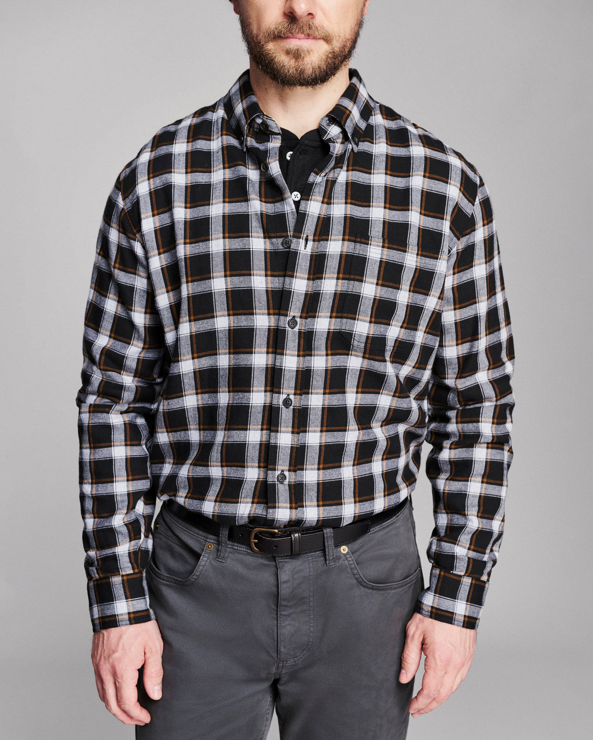 A man wearing a black, white, and thin gold plaid button-down shirt, paired with dark gray trousers and a black belt, against a solid gray studio background.