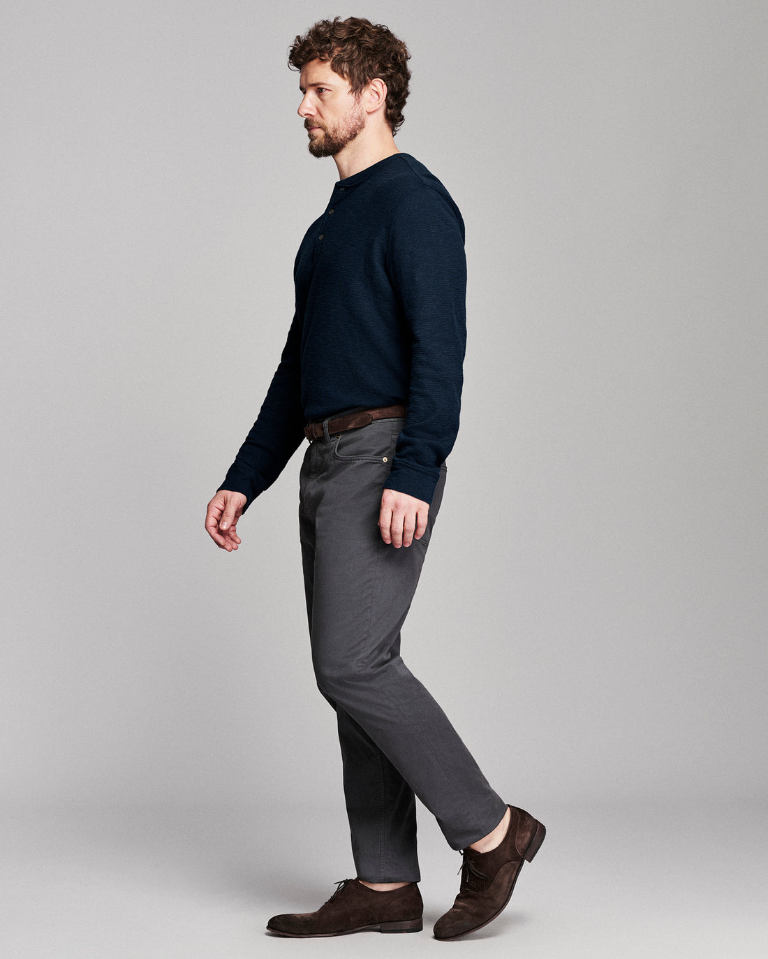 A man in a side profile wearing a dark navy long-sleeve knit sweater, charcoal gray trousers, and dark brown leather shoes, standing against a light gray studio background.