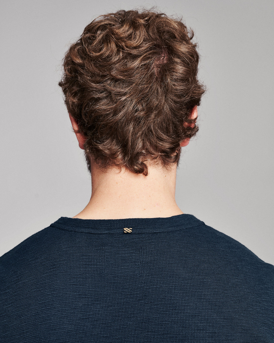 Back view of a person with curly brown hair wearing a dark navy ribbed crewneck sweater against a light gray background.
