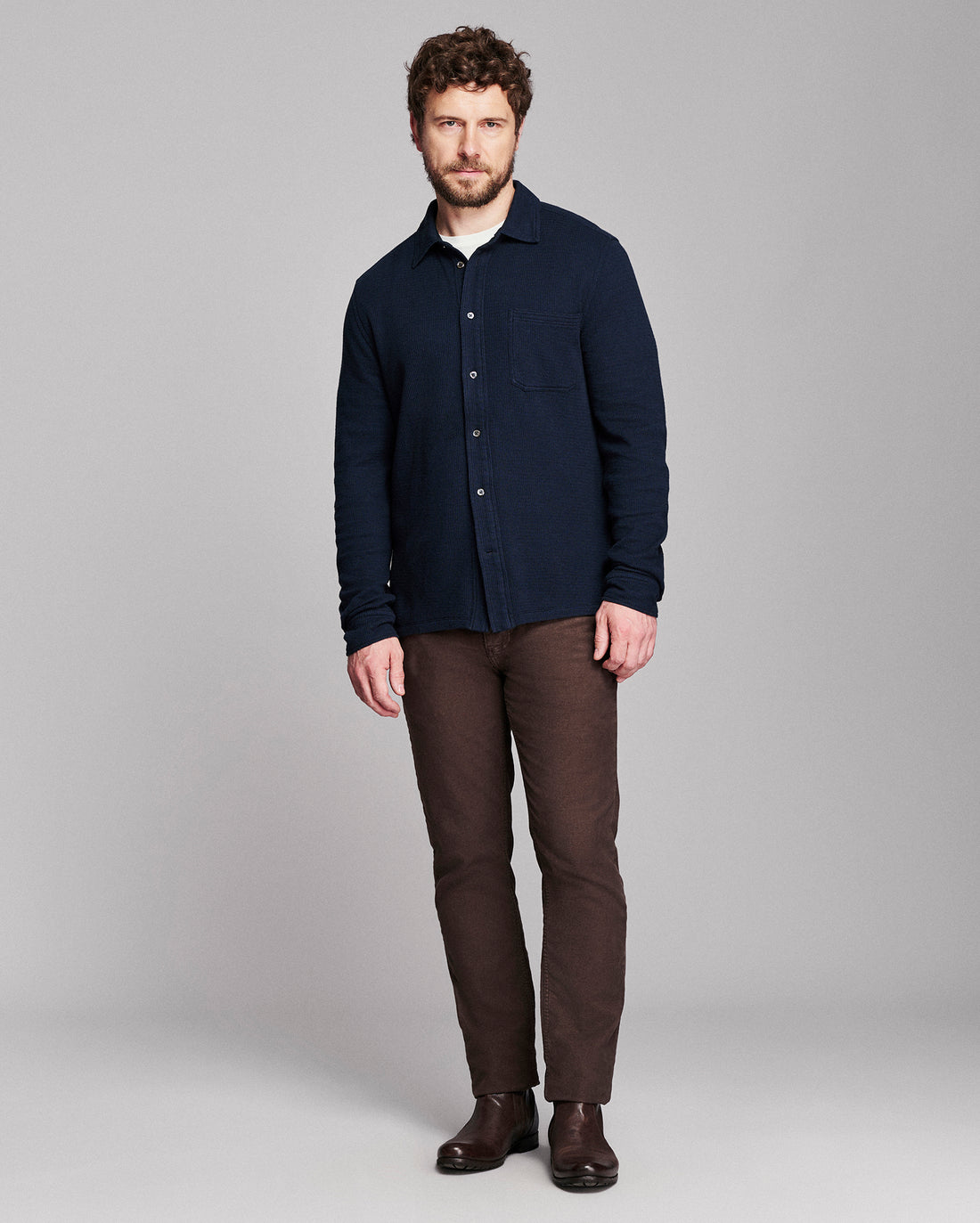 A man modeling a dark navy blue long-sleeve button-up shirt, paired with brown trousers and dark brown boots, against a solid gray studio background.