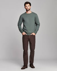 A man modeling a sage green crewneck sweater and dark brown trousers against a light gray studio background.