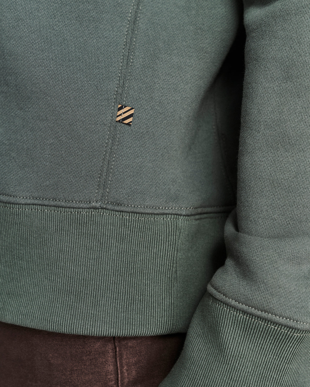 Close-up of a sage green sweatshirt showing the ribbed hem, sleeve cuff, and a small embroidered logo patch on the chest.