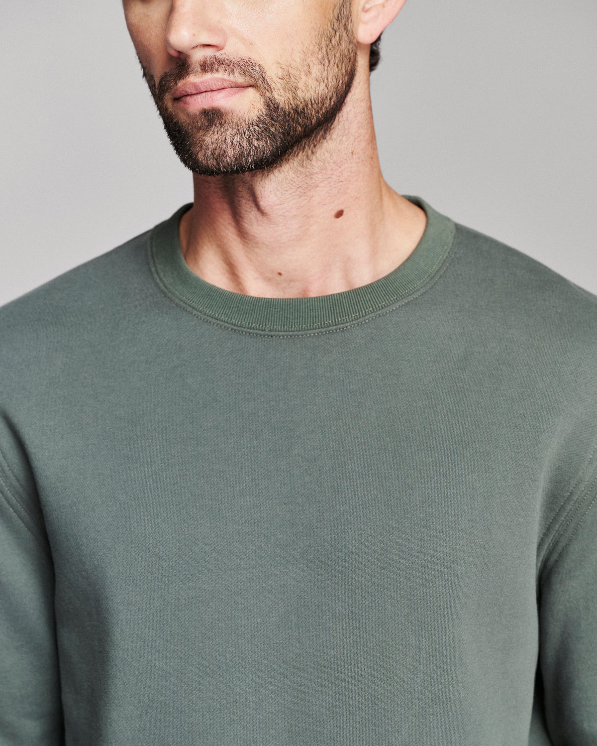 A close-up of a man wearing a muted sage green crewneck sweatshirt against a neutral gray background.